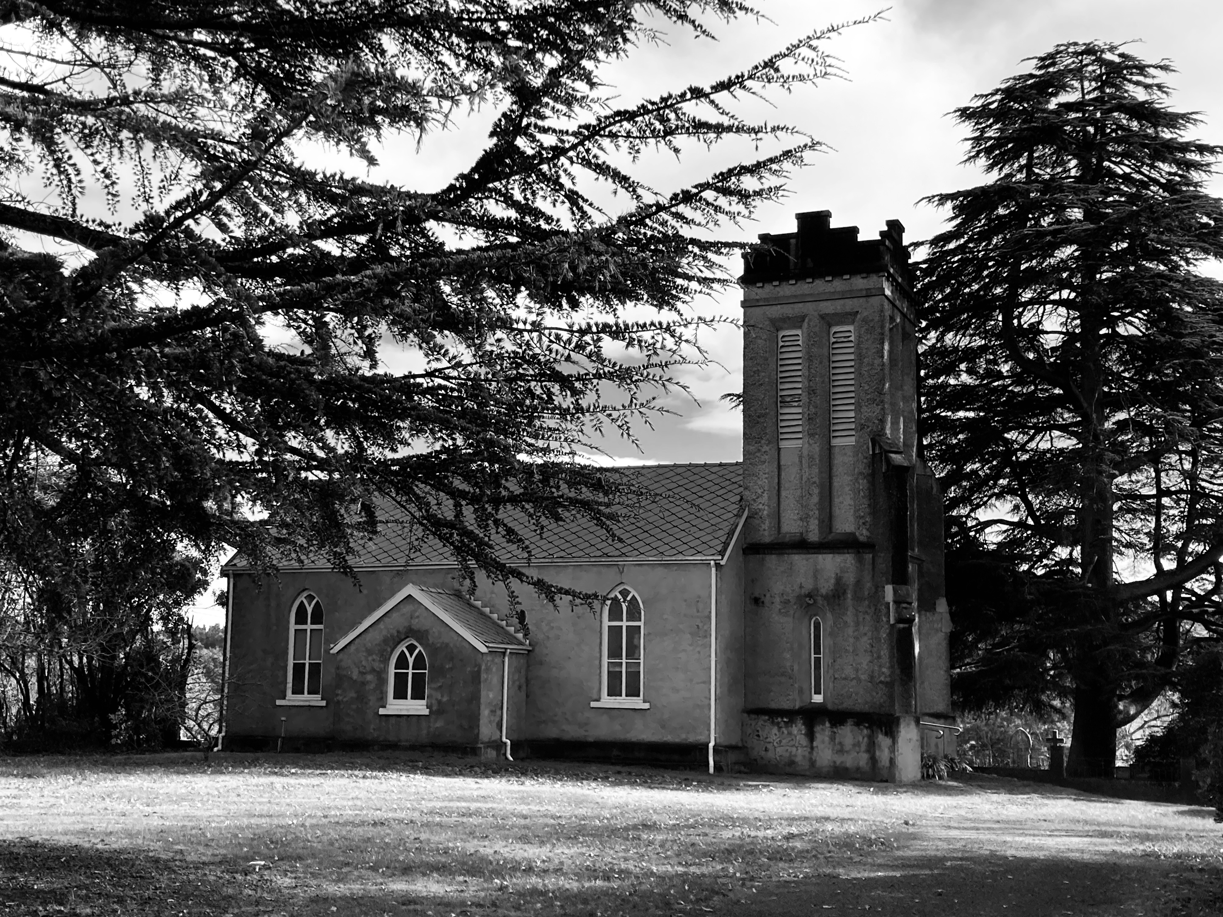 Black and white photo of a Church surrounded by trees. Graves are seen in the distance. Carrick, Tasmania.