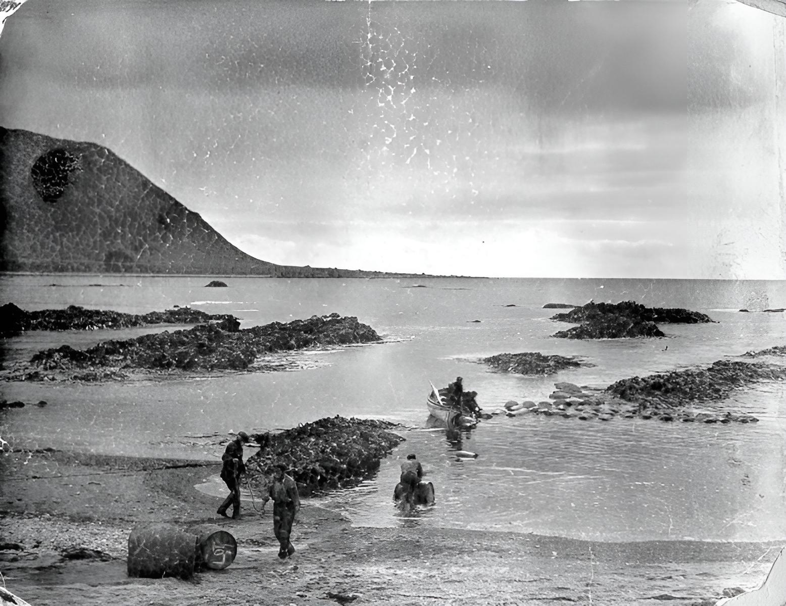 Historic photo of seamen on the rocky shoreline of Macquarie Island during the late 19th Century, unloading supplies beside the sub-Antarctic surf.