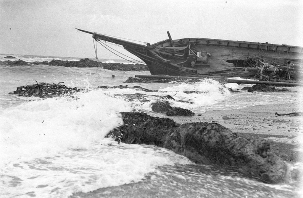 Black and white photograph shows the wrecked ship Clyde laying on it's side in the surf on the rocky shoreline of Macquarie Island.