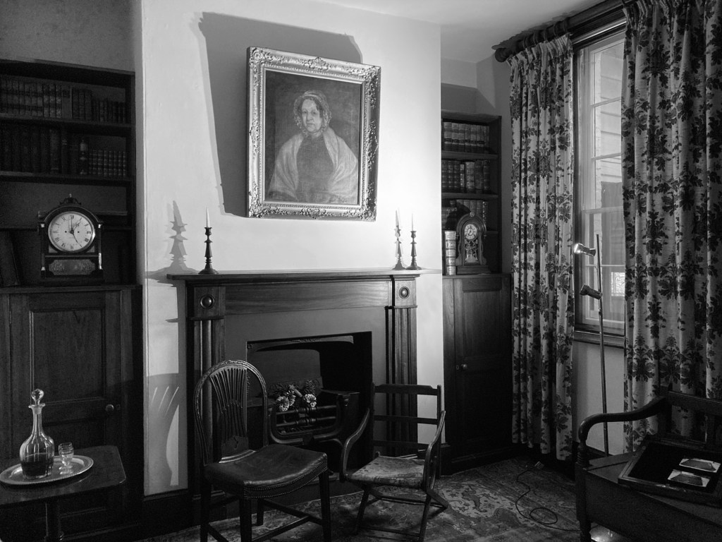 A dimly lit sitting room inside Franklin House, with a portrait above the fireplace, wooden shelves, and antique chairs, creating a quietly atmospheric historic scene.
