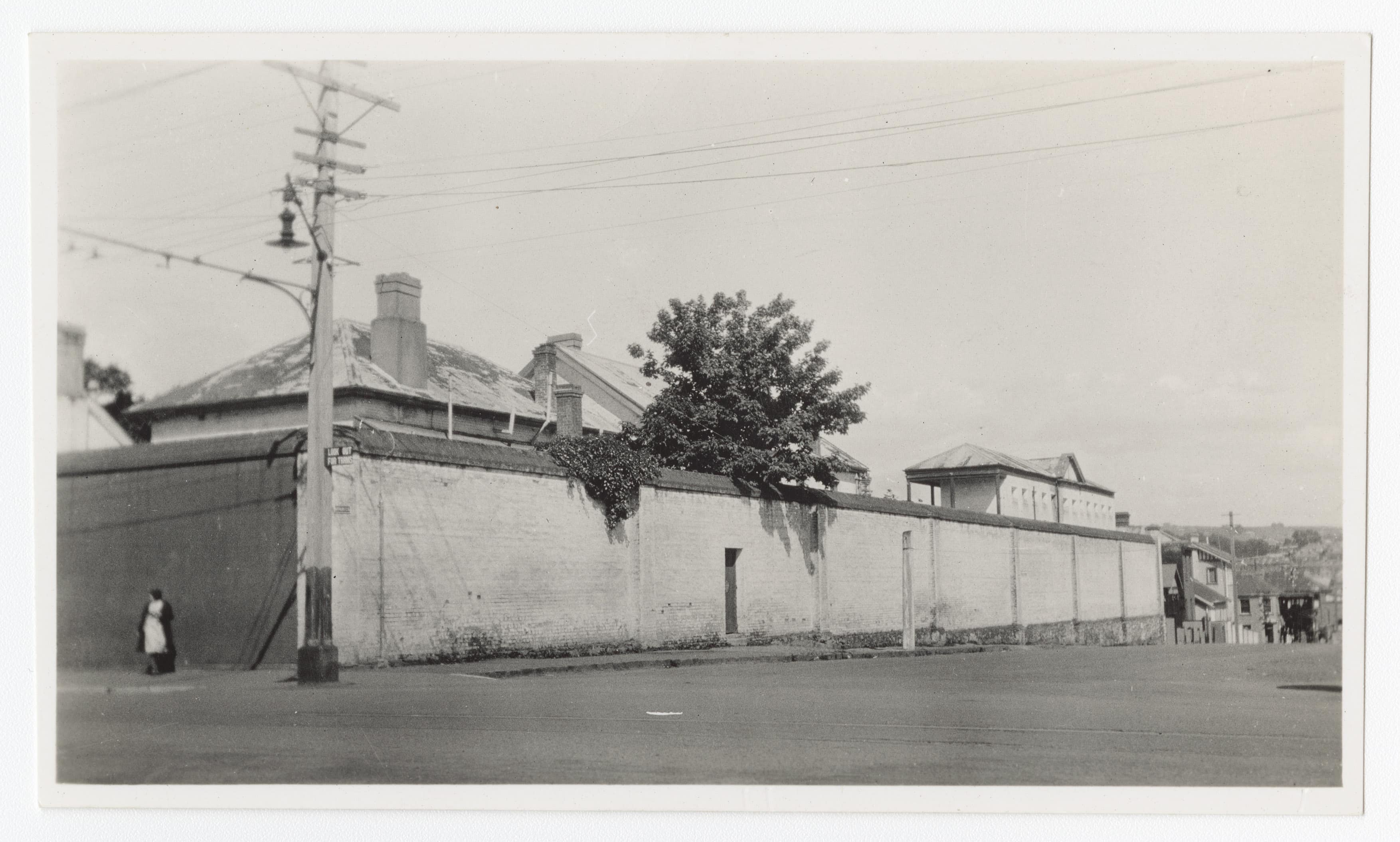 Black and white view of the Old Launceston Gaol from Bathurst Street, showing high brick walls, a pedestrian passing and early twentieth century rooftops.