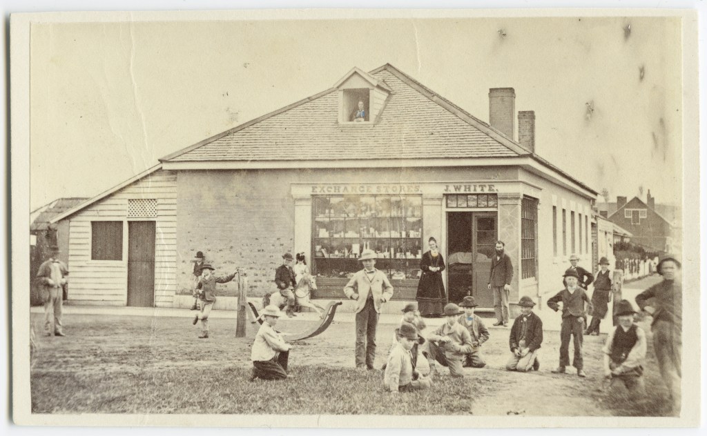 Historic photograph of children gathered outside White's Store in Bothwell, Tasmania, with boys playing in the dirt and adults standing near the shop entrance.