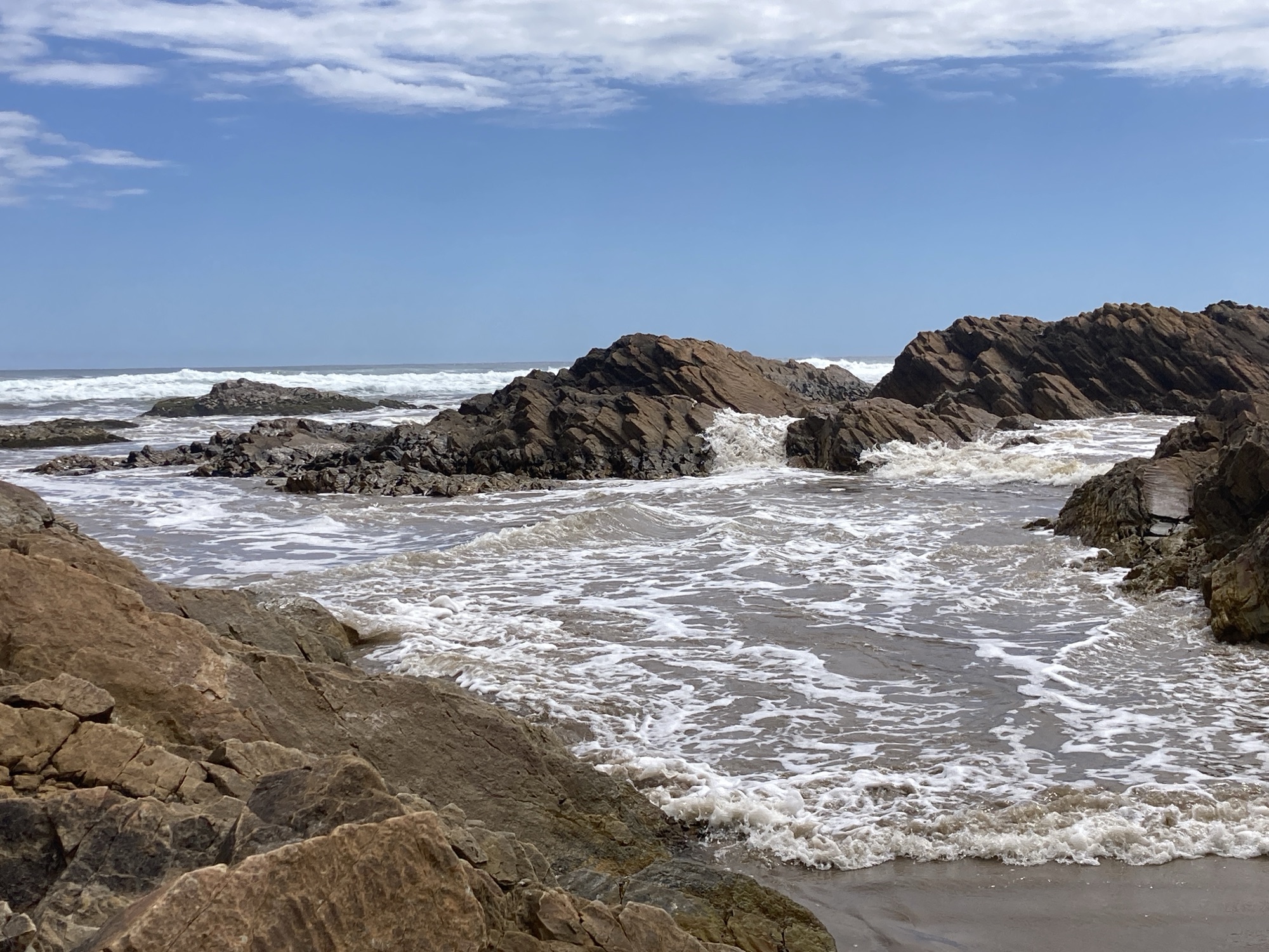 Rocky inlet on Tasmania’s west coat near Sandy Cape, with waves surging through jagged brown rocks under a bright sky.